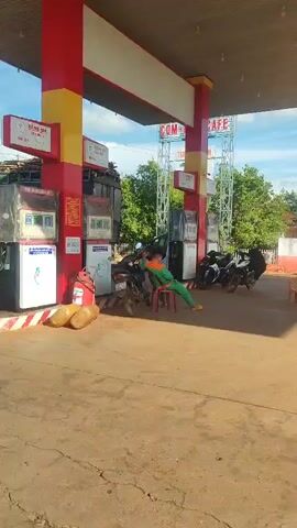 This video shows a gas station in a sunny, outdoor environment with a blue sky and scattered clouds. In the foreground, a person in an orange and green jumpsuit is sitting on a red plastic chair at a gas pump. Several motorcycles are parked around the gas station. In the background, a white and red building with yellow accents is visible, along with some trees and a few buses. The ground is paved with concrete. The camera pans across the scene, showing different angles of the gas station and its surroundings.