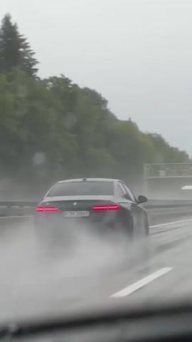 A black sedan is driving on a wet highway in the rain. The car is kicking up a lot of spray from the water on the road. There are trees and greenery on the side of the highway, and an overpass is visible in the distance. The sky is overcast and gray. The car's taillights are illuminated.