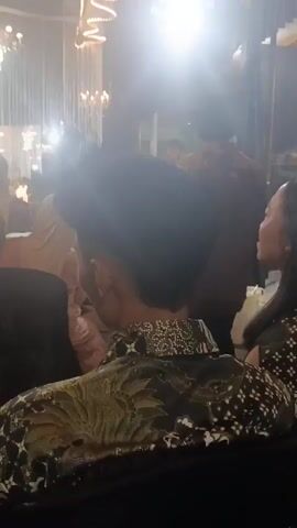The video shows a dimly lit indoor event, possibly a reception or party, with ornate lighting fixtures hanging from the ceiling. The camera is positioned behind a person wearing a patterned black shirt with gold accents. In the foreground, the back of this person's head and shoulders are visible. The camera then pans slightly to the left and focuses on a group of people. A man in a black t-shirt is holding a young child, who is wearing a dark dress and pink shoes. The man appears to be offering something on a small plate to someone off-camera. Several other individuals, mostly women dressed in modest attire including headscarves, are standing around them. The flooring appears to be artificial turf. The overall atmosphere is celebratory.
