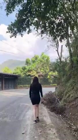 A woman in black athletic clothing walks away from the camera on a paved road.  A dark garage is on the left with foliage climbing on the wall.  Mountains are visible in the background under a blue sky with scattered clouds. Power lines and a utility pole are also visible. The road is edged with vegetation on the right side.