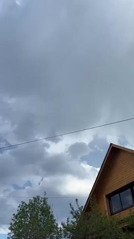The camera pans upwards from the roof of a wooden building, across a tree, wires, and a light pole, to show a sky with both gray and blue areas and white clouds.