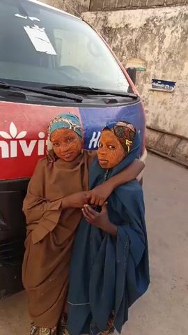 Two young individuals, presumably children, stand close together with their arms around each other in front of a van. They are wearing traditional clothing and head coverings, and their faces are covered in a brownish-orange substance. The van behind them is red and blue with a white logo. The background consists of a light-colored wall and a hint of sky.