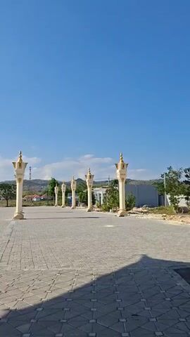 The video begins with a view of an outdoor paved area with decorative pillars lining the path. The pillars are white and gold and are topped with umbrella-like structures. In the background, there are trees, hills, and some buildings. The sky is clear and blue. As the video progresses, the camera pans to the left, revealing more of the paved area and the pillars. The camera then focuses on a large, black, cubical structure that resembles the Kaaba in Mecca. The structure is adorned with gold detailing and has a door on one side. The umbrella-like structures are visible overhead, providing shade. The scene appears to be a religious or cultural site.