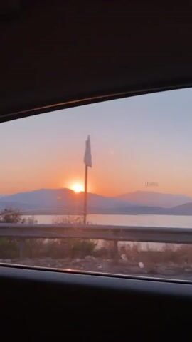 This is a short, static video shot from inside a vehicle, looking out of a side window. The sun is setting behind a range of distant, hazy mountains, casting a warm orange and pink glow across the sky. A body of water, possibly a lake or the sea, is visible in the middle ground, and beyond it, more mountains. In the foreground, the edge of the car window frame is visible, along with a metallic guardrail and some sparse vegetation. The overall impression is one of a scenic, tranquil drive during sunset.