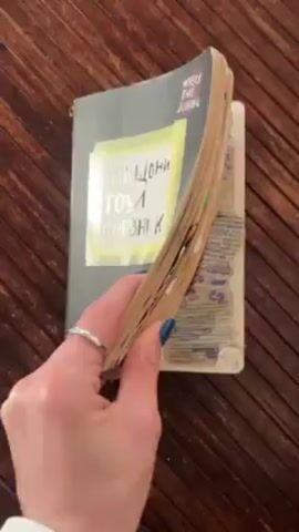 A person with blue fingernails and a silver ring on their left hand is holding a book. The book is open and they are leafing through the pages. The title of the book is "Wreck This Journal" and beneath that, in another language, is the title "Унищожи този дневник". The book has many items stuck between the pages, and the pages appear aged and filled with various marks and doodles. The book is being held over a wooden surface with a ribbed texture.
