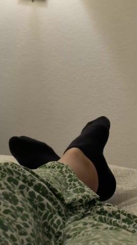 The video shows a pair of feet wearing black socks with a small white logo near the toes. The feet are lifted against a white wall, covered by a blanket with a green and white floral pattern. The lighting is soft, and there is a light fixture visible on the wall above the feet. The feet are moved slightly during the video.