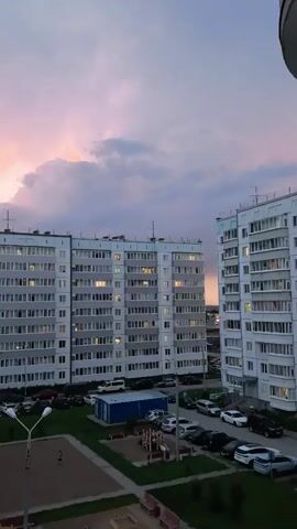 The video captures a twilight scene in Froli, Russia, focusing on two apartment buildings. The sky is filled with dramatic clouds, painted in hues of pink and purple from the setting sun. The buildings, typical of Soviet-era mass housing, are tall and rectangular, with numerous windows, many of which are illuminated, suggesting the time of day is evening.

In the foreground, a parking lot is visible with several cars parked, indicating this is a residential area. A small playground with sand and equipment is also present, hinting at families living in the buildings. A lone figure, a child, can be seen playing on the sand. The presence of streetlights suggests the area is illuminated for public safety.

The overall mood is calm and serene, amplified by the soft colors of the sky. The location cues and architectural style strongly suggest a post-Soviet urban or suburban environment. There is no visible text in the video.