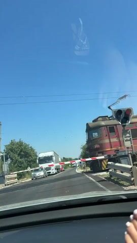 A red and yellow train with an overhead pantograph passes through a railroad crossing on a clear day. The train is moving from left to right in the frame. The crossing gates are down, and a red signal light is visible. The view is from inside a car, with the dashboard and windshield reflected in the frame. The train appears to be composed of at least two locomotives and several freight cars. The freight cars are brown and have graffiti on them. A person's hand with painted fingernails is visible in the lower right corner of the frame.