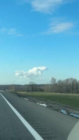 The video shows a view from inside a moving vehicle, likely a car, driving on a paved road. The sky is blue with scattered white clouds. The landscape consists of fields and trees on either side of the road. A single, bare tree stands out against the horizon. The road has a white dividing line, and some mud is visible on the side. There are no people visible in the frame. The car seems to be moving at a steady pace.