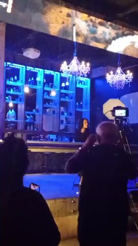 A video recording shows a woman standing behind a bar, with shelves of bottles behind her. She is speaking and gesturing. A camera on a tripod is positioned in front of her, with a person holding a phone to their ear standing to the left of the frame and another person in the foreground, partially obscuring the camera. Two chandeliers hang from the ceiling, and blue lights illuminate the shelves behind the bar. A projector is visible on the ceiling to the left. The background shows a mural or projection of a mountainous landscape.