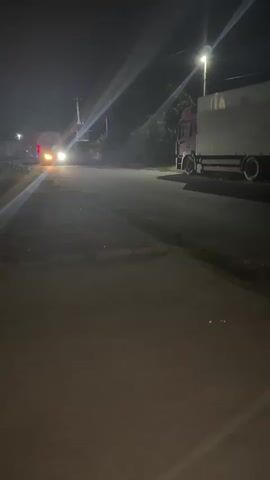 At night, two trucks are visible on a paved road. One truck, red and white, is parked on the right side of the road, facing left. Another truck, white and red, is approaching from the left side of the screen, its headlights on. The approaching truck has blue and red lights on its back. The environment appears to be a rural or semi-rural area with trees and some structures visible in the background. The road is dimly lit by a streetlamp.