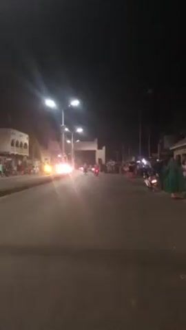 At night, a street scene is captured from a stationary, ground-level perspective. The street is paved and lit by tall streetlights, creating bright flares in the camera. Several cars and motorcycles are seen driving toward the camera, their headlights adding to the overall brightness. On either side of the street, there are buildings, people, and parked motorcycles. In the distance, an archway-like structure stands across the street. The general atmosphere suggests an active night scene with people gathered and vehicles moving along the street.