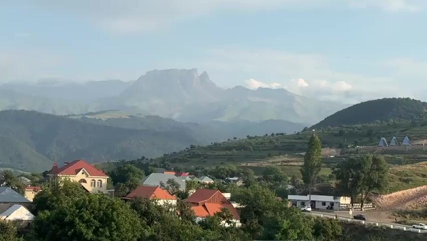 A wide shot captures a rural village nestled in a valley, with lush green hills and a prominent, layered mountain range in the background. Several houses with red and orange tiled roofs are visible in the foreground and midground, surrounded by trees. A road runs along the right side of the scene, with a few cars. The sky is a pale blue with scattered clouds. The overall atmosphere is peaceful and serene, with the natural landscape dominating the view.