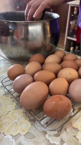 A person is dying Easter eggs. The person, wearing latex gloves, uses a spoon to place a brown egg into a series of colorful bowls. There are at least five bowls, containing dye in colors of red, blue, green, black, and yellow-orange. There are about 15 eggs on a wire rack, and a pot of water with steam is visible. The table is covered with a yellow floral patterned tablecloth.