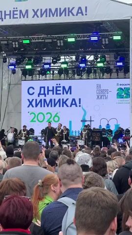 The video shows a band performing on an outdoor stage in front of a large crowd. The backdrop is a large screen displaying a message in Russian celebrating “Chemist’s Day” in 2026, along with a company logo for “PhosAgro”. The band members, dressed in formal attire with gold accents, are playing percussion instruments, including drums. The crowd, of mixed ages and backgrounds, is dense and mostly facing the stage. Many people are holding up their phones, presumably recording the performance. The environment is an outdoor venue with professional stage lighting and sound equipment.
