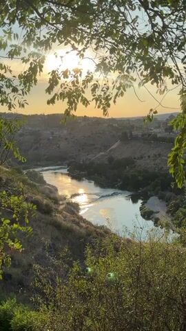 This is a landscape video shot from a high vantage point overlooking a river valley. The sun is setting, casting a warm glow on the scene. In the foreground, branches and leaves of trees frame the view. The river is visible in the middle ground, reflecting the sunlight. Beyond the river, a town or city is nestled in the hills. The overall mood is peaceful and serene.