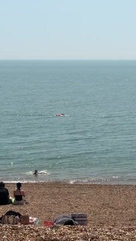 The video shows a pebble beach with two women sitting near the water's edge. One woman is wearing a black tank top. In the distance, two people are swimming in the water. One of the swimmers is using a pink flotation device. The water appears blue and is slightly wavy. The sky is a clear, pale blue.  There are several items on the beach near the women, including a patterned box, a black jacket, and a tote bag.