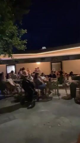 The video shows an outdoor seating area at night, possibly a restaurant or cafe. A group of people are seated at tables, some appearing to be dining or socializing. There are trees with foliage casting shadows on the ground. A stroller is visible in the foreground. Lights are illuminating the area, and some neon lights are visible in the background. The overall atmosphere appears to be relaxed and casual.