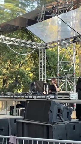 A woman stands behind a DJ booth on an outdoor stage, operating turntables and a mixer. The DJ booth is lined with multiple speakers and equipment. The stage is decorated with a mirrored surface overhead. In the background, trees and foliage are visible. A blue light is visible on a truss structure behind the DJ. The scene appears to be a live music performance.