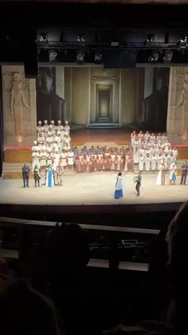 Here's a description of the scene in the video:

The video shows a scene from a staged performance, likely an opera or musical, taking place on a large stage in a theater. The audience is visible in the foreground, with the main focus on the stage.

The stage backdrop is a digitally projected image of an Egyptian-style temple or corridor, flanked by large statue images. On the stage, there is a large cast of performers. They are wearing various costumes, some in white robes and headdresses, others in simple attire suggesting servants or slaves, and a few dressed in more elaborate, regal clothing.

The performers are arranged in tiered rows. There are two performers in the forefront, one dressed in blue and white regal attire who has received a wreath from another performer. The rest of the cast is arranged behind them.

The audience is applauding, likely at the conclusion of a scene or performance.
