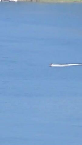 The video shows a jet ski moving across a large body of water. The water is blue, and there are mountains and trees in the background. The sky is also blue. The video is shot from a distance, and the jet ski is small in the frame. The jet ski is creating a wake behind it, indicating that it is moving at a good speed. The water appears calm.
