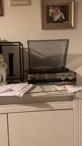 A record player is on a shelf, its lid open. A record is on the turntable and is spinning. The record player has several buttons and knobs on its front. There is a framed picture on the wall above the record player. To the left of the record player is a black shelf unit with a statue on it. Papers are scattered on the shelf in front of the record player.