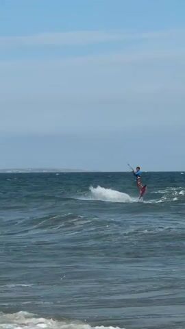 A kitesurfer in a blue shirt and dark shorts is performing a jump over the ocean. The kitesurfer is wearing a red board and is in mid-air, facing left. The ocean has moderate waves and a clear blue sky with some clouds is above. In the distance, land with some buildings and a lighthouse is visible. Another kitesurfer with a green kite is further in the distance. Several people are swimming in the water closer to the shore.