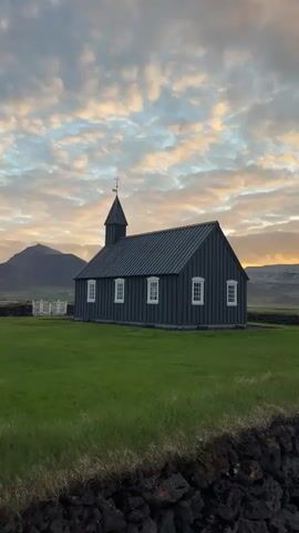 The video captures a static, eye-level view of the iconic black wooden church, Búðakirkja, in Búðir, Iceland. The church, painted a dark charcoal grey or black, stands prominently in a lush green grassy field. It features a traditional steeple topped with a cross and a weather vane, and several white-framed windows on its side. A small white fence, likely part of a churchyard or memorial plot, is visible behind the structure.

In the immediate foreground, a dark, rugged stone wall runs across the bottom of the frame, providing textural contrast. The background reveals rolling mountains under a dramatic sky filled with clouds. The clouds are illuminated with soft, warm hues of orange, pink, and yellow, suggesting either sunrise or sunset. The light creates an ethereal glow over the landscape, enhancing the serene and tranquil atmosphere.

On the bottom right, a small sign is partially visible behind the stone wall. It depicts silhouettes of figures and text, which appears to include "Minningarreitur" (memorial plot or graveyard) and potentially "Búðakirkja" below it. No people are visible, and no activity is taking place, allowing the viewer to focus on the architectural beauty of the church against its stunning natural surroundings. The scene evokes a sense of peaceful solitude and the distinctive natural beauty of Iceland.