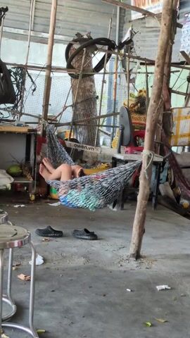 Here is a concise description of the video:

The scene captures a person lying in a hammock, possibly indoors on a cement floor. Two black pairs of slip-on shoes sit near the base of one of the hammock’s wooden supports. Various objects and furniture are scattered around the room, including a metal stool in the foreground, a cabinet, boxes, and what appears to be a fan in the background. The room seems cluttered and has a somewhat makeshift quality to it. The hammock itself is made of a netting material, and the person appears to be looking at a mobile phone.