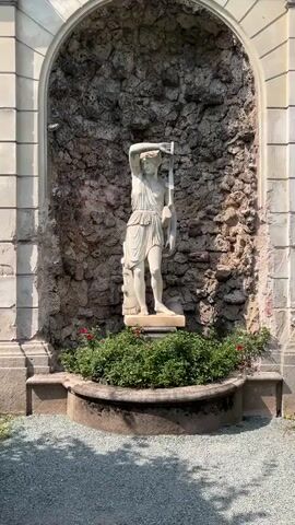 A white marble statue of a standing male figure is positioned in a stone alcove. The figure is depicted with a chiton and is holding a spear vertically with his right hand raised. In front of the statue, a circular planter is filled with dense green foliage and a few small red flowers. The alcove's background is a rough, textured stone wall. Sunlight casts shadows on the ground in the foreground.