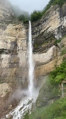 The video shows a tall, thin waterfall cascading down a sheer, layered rock cliff face. The rock is a mix of tan, brown, and gray, with dark horizontal striations. Lush green vegetation clings to the sides of the cliff and grows at the base of the waterfall, where the water breaks into smaller streams and pools over rocks. The top of the waterfall is shrouded in mist or fog, obscuring the origin of the water and the higher parts of the cliff. The overall atmosphere is one of natural grandeur and slight mystery due to the obscured upper portion.