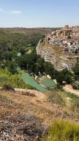 The video shows a high-angle view of a river winding through a valley. On the right side of the valley, a town is built into the side of a rocky cliff. The town consists of white and terracotta-colored buildings, with a prominent church spire visible. Lush green trees line the riverbanks and cover the hillsides. The river itself is a turquoise green color and appears to have some man-made structures, possibly dams or pools, along its course. The foreground consists of dry, sparse vegetation and rocks. The sky is clear and blue, indicating a sunny day. The camera appears to be stationary or panning very slowly, capturing a wide panorama of the landscape.
