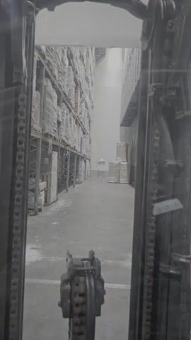 The video shows a view from inside a forklift, looking down a warehouse aisle. Tall shelves stocked with boxes line both sides of the aisle. Pallets of goods are stacked on the right side of the aisle. The floor is concrete. The lighting is artificial, and the scene appears to be in a well-maintained industrial setting. The forklift appears to be stationary at the beginning of the video, and the camera is static.