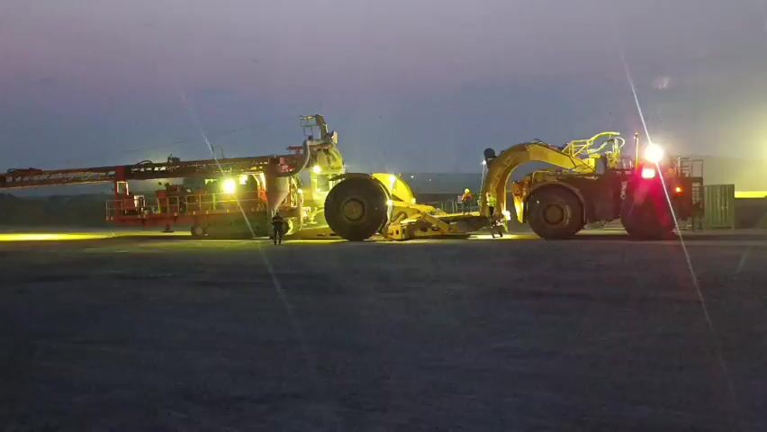 This media file shows a mining operation at dusk in Қопа ауылдық округі, Kazakhstan. The scene is dominated by heavy mining equipment, including a large articulated hauler and a drilling rig, both illuminated by artificial lights. Two figures, presumably workers, are visible near the machinery, suggesting ongoing activity. The sky is a gradient of deep purple and orange, indicative of twilight, with bright lights from the equipment casting long shadows on the dusty ground. The weather appears calm, with no visible precipitation or strong winds. The overall impression is one of industrial activity in a remote, arid environment during the transition from day to night. No text is visible in the media file.