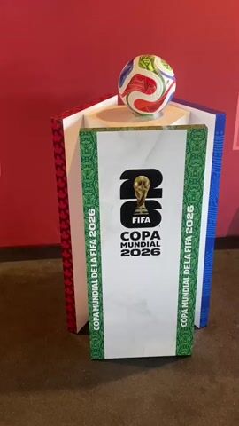 The video displays a close-up view of a FIFA World Cup 2026 display. A colorful soccer ball rests on a white pedestal, which is inside a hexagonal structure. The front of the pedestal features the "2026 FIFA COPA MUNDIAL 2026" logo. The background is a plain red wall. The camera pans slowly to capture different angles of the display, focusing on the soccer ball and the logo. There are no people visible in the video.