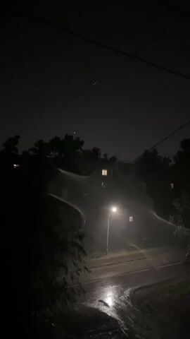 The video captures a nighttime scene, likely during a rainstorm. A street light illuminates a wet road, reflecting the light. Trees and dark shapes of houses are visible in the background, shrouded in darkness. The rain appears heavy, creating a hazy effect around the street light. No people are visible in the footage. The overall atmosphere is dark and wet.