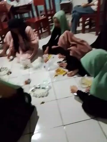 Several women are sitting on the floor of a room, eating. They are sitting on a tiled floor and appear to be eating from small white plastic bowls, with wrappers and plastic bottles strewn about. Some of the women are wearing hijabs. There are tables and chairs in the background.