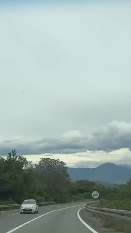 The video shows a view from inside a car driving on an asphalt road. The road curves gently and is marked with white lines.  On the left side of the road, there is a grassy area with trees, while on the right side, there is denser vegetation. In the distance, there are mountains with snow-capped peaks visible against a cloudy sky. The clouds are predominantly gray and white, creating an overcast atmosphere. A road sign warning of an upcoming blind curve is visible on the right side of the road. A white car is traveling ahead, and further down the road, a red truck and other vehicles are also visible.