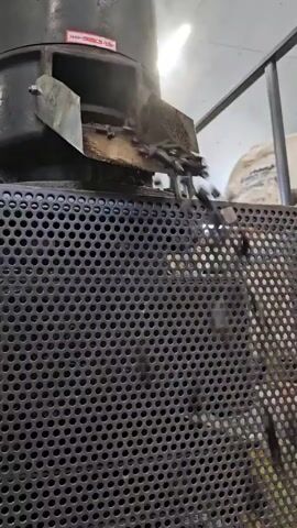 The video shows a close-up of industrial equipment, likely a pellet mill or similar processing machine. Small, dark pellets are being ejected from a chute onto a perforated metal grate. The pellets appear to be falling freely, suggesting they have just been formed or are being discharged. The machine is dark-colored, with some signs of wear and dust. The background is blurred but appears to be an indoor industrial setting with lighting fixtures visible. No people are visible in the frame.