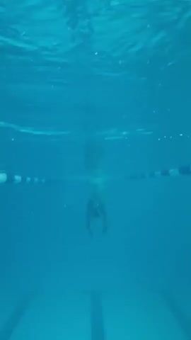 An underwater shot in a swimming pool shows a person swimming. The person is wearing blue swim trunks and a swim cap, and is swimming towards the camera. The water is a clear blue, and the bottom of the pool is visible, with lane lines marked on it. The surface of the water is visible at the top of the frame, with ripples and light reflecting off it.