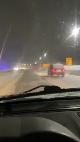 The video, shot from the interior of a car, shows a snowy road at night. The windshield wipers are active, clearing snow and moisture from the glass. The road is illuminated by streetlights, creating a wet and reflective surface. Several vehicles can be seen in the distance, their tail lights glowing red. One vehicle, a small dark-colored car, is closer and visible on the right side of the road. The overall environment suggests a winter night with challenging driving conditions due to snow and low visibility.