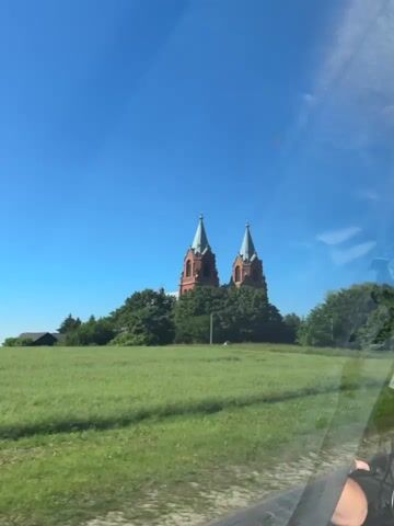 The video shows a church with two spires, made of brick, against a bright blue sky. The church is surrounded by trees and a grassy field. The camera appears to be moving, possibly from inside a vehicle, as evidenced by the reflections on the right side of the frame and the slight blurriness. A person's arm is visible in the lower right corner of the frame. The overall impression is of a sunny day and a rural landscape.