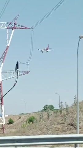 The video shows a close-up view of a large, red and white lattice tower, likely a high-voltage electricity pylon, against a clear blue sky. The tower has multiple crossbars and insulators, with thick cables suspended from it. The camera appears to be moving, as evidenced by the slight blur in the background, suggesting it might be from a vehicle. Trees and bushes are visible at the base of the tower. The video then transitions to a blurred shot of a concrete structure, possibly a bridge or overpass, with graffiti on its underside. A section of a metal railing is also visible in the foreground, and further motion blur indicates continued movement. The final frames are overexposed and feature a bright white glare, obscuring any discernible details.