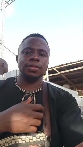 This media file is a short video with a clear focus on a young Black man taking a selfie. He is wearing a black tunic with intricate gold embroidery around the neckline and down the front. A brown strap from a bag is visible across his chest. His hair is short and fades on the sides. He has a medium complexion and a slight smile. In the background, there appears to be an outdoor market or gathering. Structures with thatched roofs and wooden beams are visible. Other people are present in the background, though they are not in sharp focus. One person in a bright yellow shirt and a headscarf with a red and green circular pattern is particularly noticeable. The lighting suggests it is daytime, likely under a bright sky. The overall atmosphere seems casual and perhaps convivial. There is no visible text in the media file. The location context provided is Wamba, Nigeria.