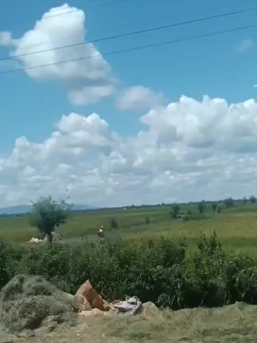 The video shows a rural landscape on a sunny day with puffy white clouds in a blue sky. The landscape consists of fields of green vegetation. Power lines run across the top of the frame. Two people walk through the field. A white truck passes by, obscuring the view.
