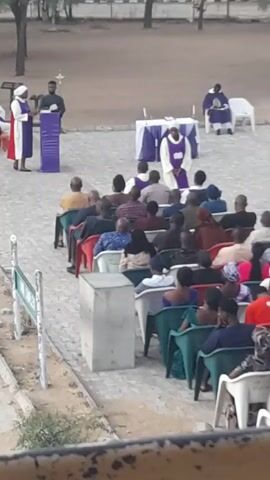Here is a detailed description of the video:

The video shows an outdoor gathering, likely a church service or a similar religious event. A group of people are seated in rows of chairs facing a small stage or podium.

At the front, near the stage, are several individuals in religious robes and attire. One person is standing at a podium, possibly giving a speech or sermon. Another person in robes stands nearby.

The audience is diverse in terms of age and attire. Most people are seated and facing the stage, listening attentively. The setting appears to be a paved area with some trees in the background.

There are also tables draped with white cloths and purple accents, suggesting an altar or ceremonial area. A loudspeaker sits a distance away from the gathering.

Overall, the scene captures a religious gathering taking place outdoors, with people engaged in a formal service or ceremony.