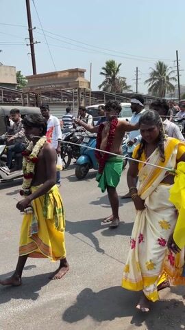 People are walking on a street, seemingly as part of a religious procession. Several people are dressed in traditional Indian attire. One individual has what appears to be a skewer piercing their cheeks. Others carry flags and decorative items, and some are wearing garlands of flowers. The environment is urban, with buildings and vehicles visible in the background.