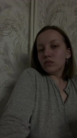A young woman with short, light brown hair is shown from the chest up, against a wall with gray and white leaf-patterned wallpaper. She is wearing a gray button-up shirt and has jewelry in her ear. She is looking at the camera, exhaling vapor, then lifts a clear glass filled with a red liquid, and drinks from it.