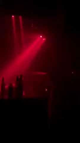 The video depicts a dimly lit nightclub atmosphere, likely during an event. Red stage lighting illuminates figures silhouetted within the crowd. A DJ booth, indicated by a dark rectangular shape, sits in the foreground.