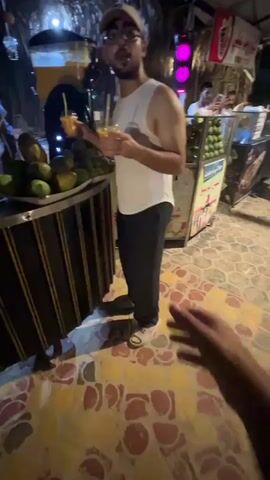 The video shows a person holding a plastic cup of orange juice with a straw. In the background, another person in a white tank top and glasses is holding two similar cups. There are several green mangoes on a tray in front of them, and a large dispenser of orange liquid is visible. The environment appears to be an outdoor market stall at night, with string lights and other vendors in the background. The person holding the cup is walking forward, and the camera is held from their perspective.